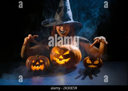 Eine ominöse Hexe im Hut zaubert über eine Jack-o-Laterne. Traditionelle halloween-Figuren. Mystischer Nebel kriecht über Kürbisse mit geschnitzten schrecklich Stockfoto