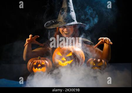 Eine ominöse Hexe im Hut zaubert über eine Jack-o-Laterne. Traditionelle halloween-Figuren. Mystischer Nebel kriecht über Kürbisse mit geschnitzten schrecklich Stockfoto