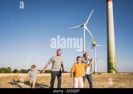 Optimistische Familie, die im Windpark läuft, Tochter, die ein buntes Windrad trägt Stockfoto