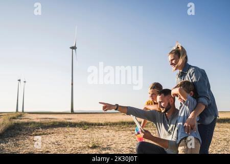 Familie mit digitalem Tablet im Windpark, der auf Windenergieanlagen zeigt Stockfoto