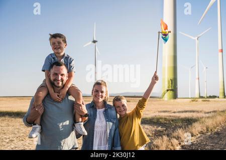 Optimistische Familie, die im Windpark steht, Tochter, die ein buntes Windrad trägt Stockfoto