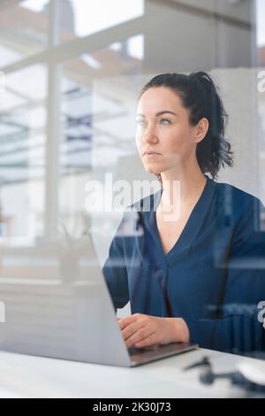 Durchdachte Geschäftsfrau, die mit einem Laptop durch das Glas gesehen sitzt Stockfoto