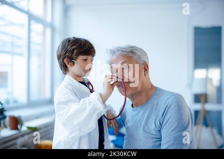 Junge imitiert als Arzt, der zu Hause die Nase des Großvaters mit Stethoskop überprüft Stockfoto