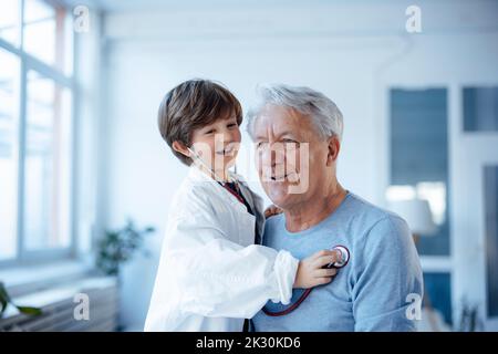 Glücklicher Junge imitiert als Arzt, der Großvater mit Stethoskop zu Hause überprüft Stockfoto