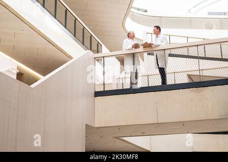 Lächelnde Mitarbeiter des Gesundheitswesens diskutieren miteinander, die in der Nähe von Geländer im Krankenhausflur stehen Stockfoto