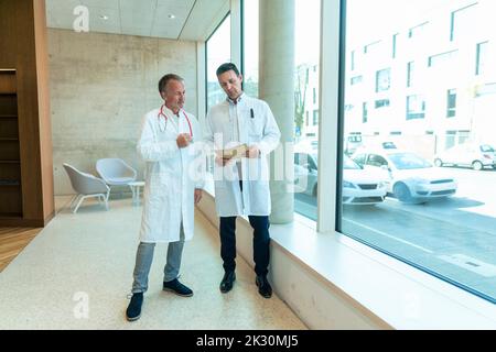 Ärzte in Laborkittel diskutieren über Krankenakte, die im Krankenhaus am Fenster stehen Stockfoto