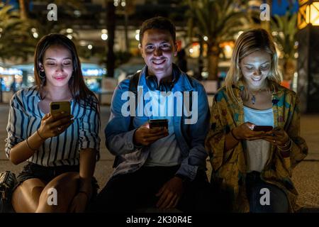 Lächelnder junger Mann, der nachts unter Frauen mit dem Smartphone sitzt Stockfoto