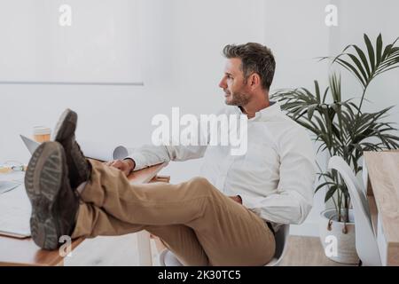 Nachdenklicher Geschäftsmann, der im Büro mit gekreuzten Beinen am Knöchel sitzt Stockfoto