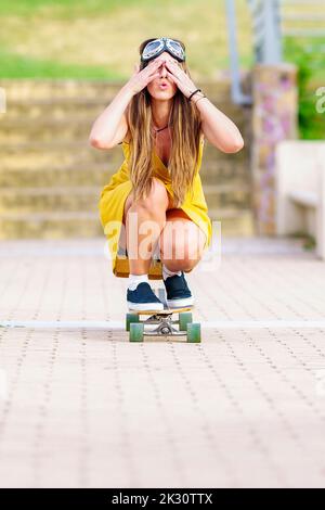 Junge Frau, die die Augen mit der Hand bedeckt und Skateboarding auf dem Fußweg Stockfoto