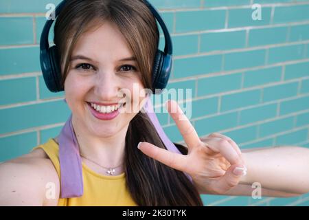 Junge Frau mit Kopfhörern, die an der Wand ein Friedenszeichen zeigt Stockfoto