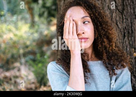 Junge Frau bedeckt das Gesicht mit der Hand, die sich am Baumstamm im Park lehnt Stockfoto