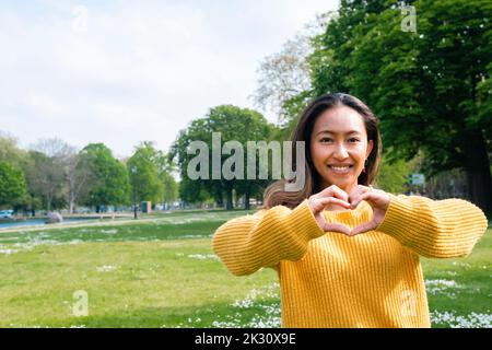 Glückliche junge Frau macht Herzform mit Händen im Park Stockfoto