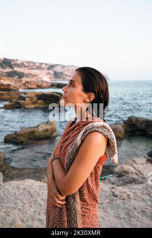 Nachdenkliche Frau mit gekreuzten Armen, vom Meer in eine Decke gehüllt Stockfoto