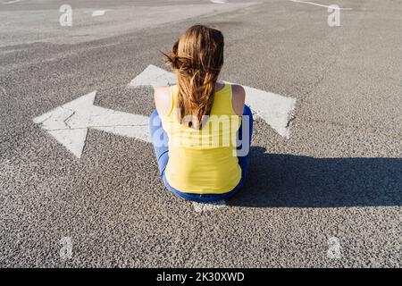 Frau sitzt auf Asphalt mit drei-Wege-Pfeil-Zeichen Stockfoto