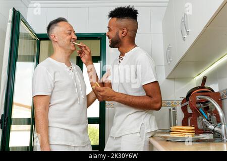 Glücklicher junger Mann füttert Pfannkuchen zu Freund in der Küche zu Hause Stockfoto