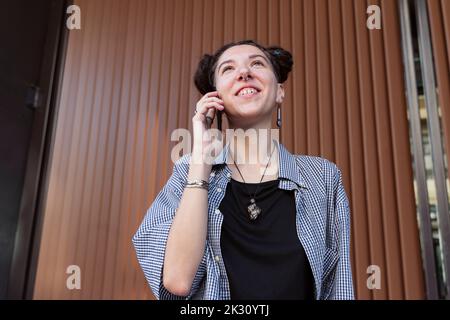 Lächelnde, nicht-binäre Person, die vor der Wand auf dem Smartphone spricht Stockfoto