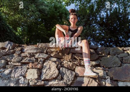 Junger nicht-binärer Hipster, der auf einer Steinwand sitzt Stockfoto