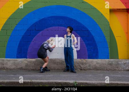 Junger Mann, der Overalls trägt, steht neben einem Freund und schreibt auf einem Regenbogen-Wandbild Stockfoto