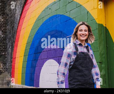 Fröhliche junge Frau, die an der Wand durch ein Regenbogengemälde läuft Stockfoto