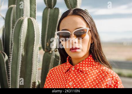 Selbstbewusste junge Frau mit Sonnenbrille, die vor einer Kaktuspflanze steht Stockfoto