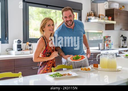 Lächelnde reife Frau mit Mann, der zu Hause am Esstisch in der Küche steht Stockfoto