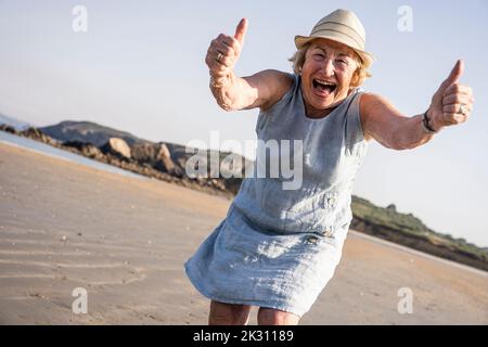 Fröhliche ältere Frau, die Daumen hoch zeigt und Spaß am Strand hat Stockfoto