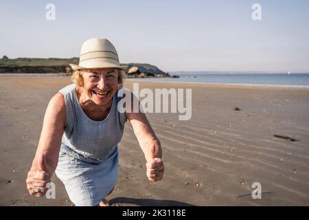 Glückliche ältere Frau, die am sonnigen Tag am Strand Daumen nach oben zeigt Stockfoto