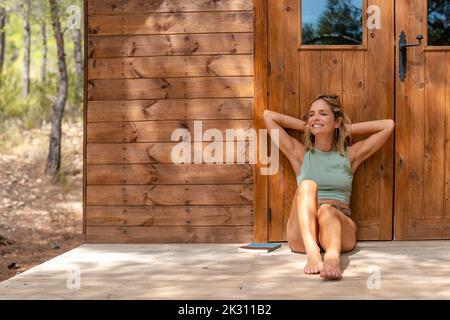 Glückliche reife Frau mit Händen hinter dem Kopf, die an der Tür auf der Veranda gelehnt sind Stockfoto