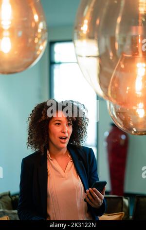 Überraschte Geschäftsfrau mit Smartphone im beleuchteten Büro Stockfoto