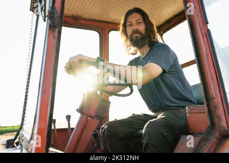 Lächelnder, reifer Bauer, der an einem sonnigen Tag im Traktor sitzt Stockfoto