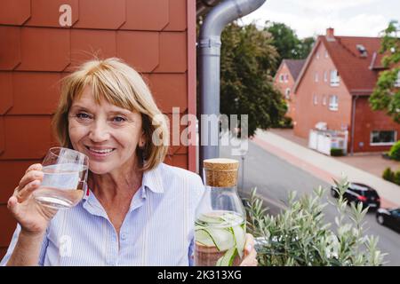 Glückliche ältere Frau mit blonden Haaren, die ein Glas Wasser trinkt Stockfoto