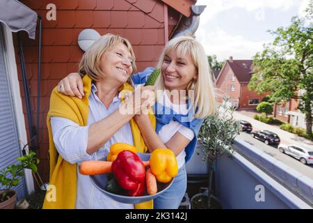 Glückliche Frau mit Mutter, die eine Schüssel mit frischem Gemüse auf dem Balkon hält Stockfoto