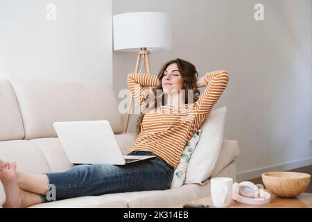 Junge Frau mit Laptop, die sich zu Hause auf dem Sofa entspannt Stockfoto