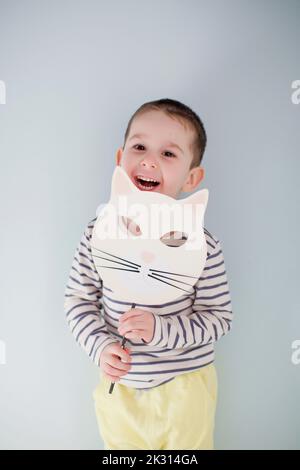 Fröhlicher niedlicher Junge, der mit handgefertigter Halloween Maske vor einer grauen Wand steht Stockfoto