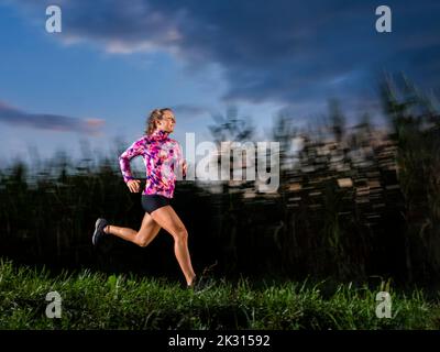 Junge Sportlerin, die in der Dämmerung auf Gras läuft Stockfoto