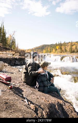 Vater und Sohn sitzen neben dem Fluss mit Angelruten