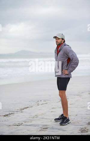 Einen morgendlichen Lauf am Strand machen. Ganzkörperaufnahme eines hübschen jungen Mannes, der an einem Strand steht. Stockfoto