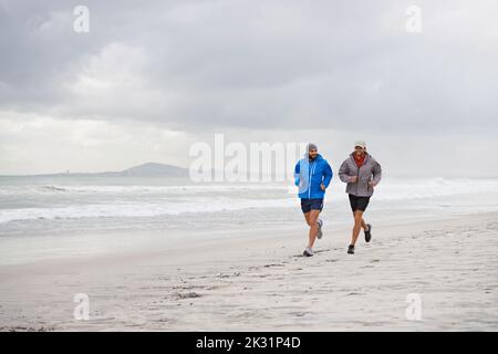 Morgens mit einem Freund am Strand laufen. Die ganze Länge der Aufnahme von zwei Männern, die an einem bewölkten Morgen gemeinsam am Strand joggen. Stockfoto