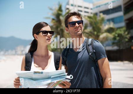 Es gibt so viel zu tun - wo man zuerst hingeht. Ein junges Paar, das auf einer Karte steht, während es am Strand steht. Stockfoto