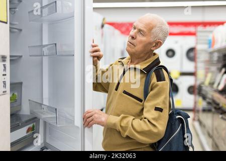 Älterer grauhaariger Rentner, der am Schalter im Ausstellungsraum der Abteilung für Elektrogeräte einen Kühlschrank sah Stockfoto