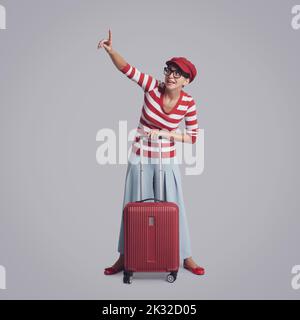 Lächelnde, aufgeregt Reisende Frau, die ihren Koffer hält und den Finger nach oben zeigt, Reise- und Tourismuskonzept Stockfoto