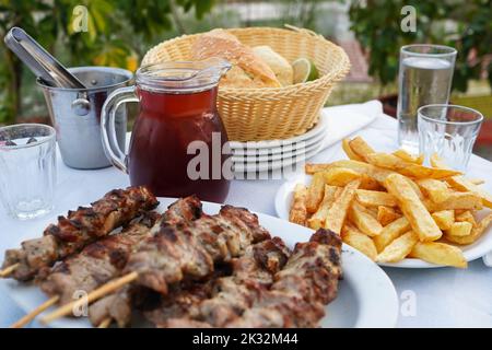 Selektiver Fokus auf Wein mit typischer griechischer Tischanordnung, einschließlich des halben Liter-Kännchens Rotwein, gebratener Kartoffeln, Souvlaki und Brot Stockfoto