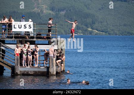 Die Menschen genießen den Sommer in Luss am Loch Lomond, Schottland, beim Springen und Schwimmen im kalten, frischen Wasser des loch. Stockfoto