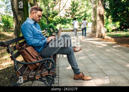 Konzentrierter erwachsener Mann, der auf seinem Laptop tippt, im Park arbeitet, seinen Job ernst nimmt. Stockfoto