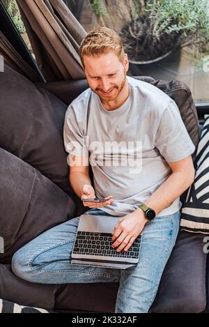 Ruhiger erwachsener bärtiger Mann, der sich zu Hause entspannt, auf seinem Laptop tippt und Telefonnachrichten überprüft. Stockfoto