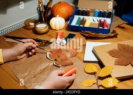 Herbsthandwerk aus getrockneten Blättern. DIY Herbst Crafts für Erwachsene. Collage aus Herbstnatur. Weibliche Hände zeichnen Fuchsgesicht auf trockenem Ahornblatt Stockfoto