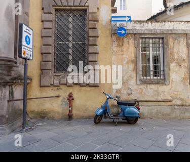 Blaue Vespa und ein gelbes Gebäude mit großen Fenstern in einer Einbahnstraße in der Stadt Lucca, Toskana, Italien Stockfoto