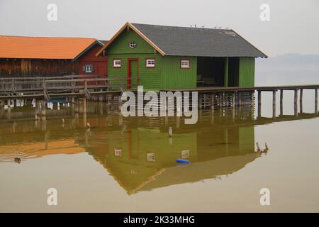 Lange hölzerne Seebrücke, die an trüben und nebligen Tagen zu den bunten Bootshäusern am Ammersee im bayrischen Dorf Schondorf führt (Bayern, Deutschland) Stockfoto