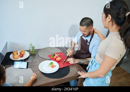 Asiatische Frau und Mädchen schauen auf Mann, der Gurke schneidet Stockfoto