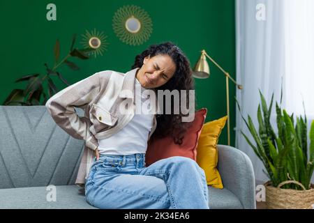 Junge schöne hispanische Frau zu Hause mit starken Rückenschmerzen, Hausfrau sitzt auf dem Sofa im Wohnzimmer und massiert Rückseite im Zimmer mit grüner Wand. Stockfoto